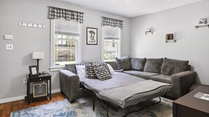 A modern living room with a grey sofa, a small table, and a bench. There are two windows with checkered curtains, a wall-mounted shelf with books, and a ceiling light.
