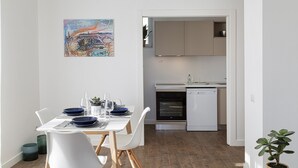 Een moderne keuken met witte kastjes, een vaatwasser en een oven. Er is een zithoek met een tafel gedekt voor twee personen, witte stoelen en een schilderij aan de muur.