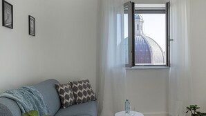 Een kamer met een grijze bank, een klein wit tafeltje met een fles en glas, een raam met uitzicht op een koepelvormig gebouw, en een plant in een geweven pot.