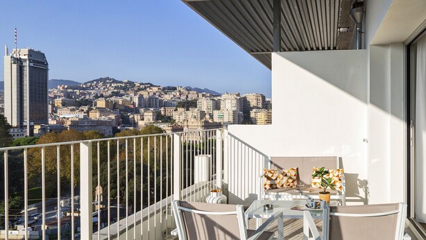 A balcony with a table and chairs, overlooking a cityscape with buildings and a hill in the distance. - Deluxe Apartment - Ice by Wonderful Italy (Genova)
