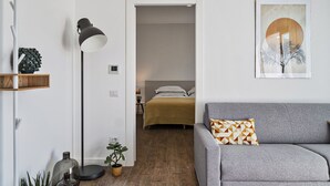 A modern living room with a grey sofa, a floor lamp, and a small table with a plant. A doorway leads to a bedroom with a bed and a nightstand. - Deluxe Apartment - Ice by Wonderful Italy (Genova)