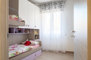 Ein Kinderzimmer mit Etagenbetten, Regalen voller Spielzeug und einem Fenster mit durchsichtigen Vorhängen.