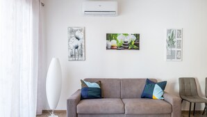 Un salon avec un canapé marron, deux coussins, un lampadaire blanc et trois œuvres d’art accrochées au mur.
