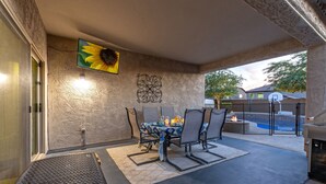 Un patio exterior con una mesa, sillas, un televisor montado en la pared y un aro de baloncesto.