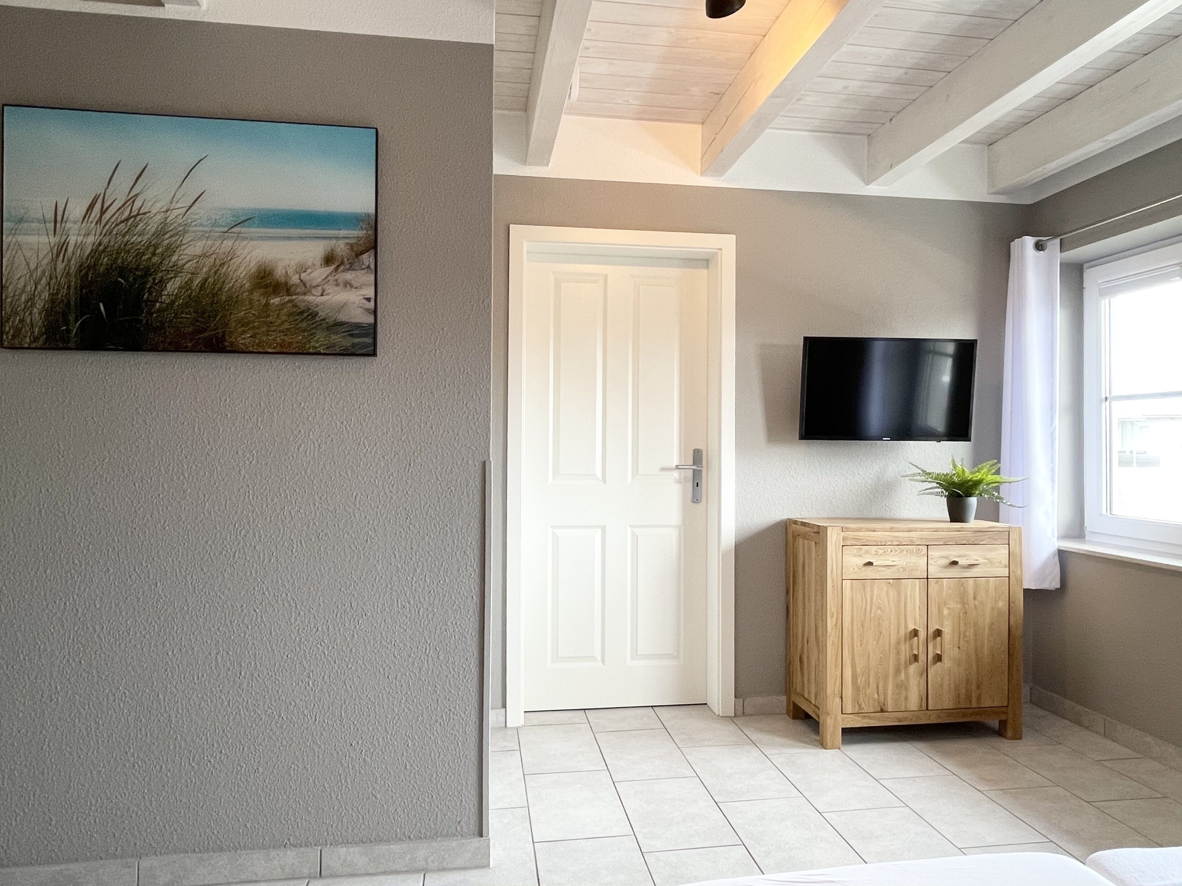 A hallway with a door, a wooden cabinet, a mounted TV, and a beach painting.