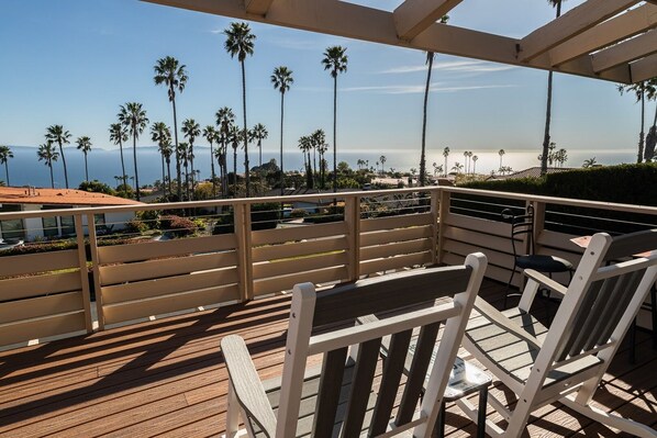 A wooden deck with white chairs and a railing overlooking a beach with palm trees and a view of the ocean.