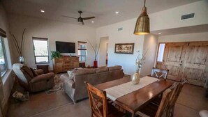 A spacious living area with a large sofa, a dining table with chairs, a ceiling fan, and a TV mounted on the wall. - 5yr old home , private, 1-BdRm home on its own 3.3 acres with city views (Tucson)