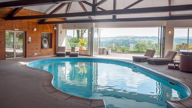 Una piscina interior con vistas al campo, vigas de madera en el techo y zona de estar exterior.