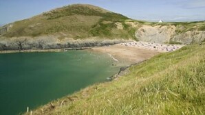 Un paisaje costero con playa, colina cubierta de hierba y una pequeña construcción blanca.