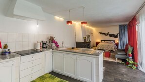 A compact kitchen with white cabinets, a coffee maker, and a small island. A bed with a red headboard and a gray sofa is visible in the background.