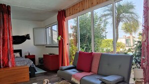 A cozy living room with a grey sofa, red and white cushions, a wooden coffee table, and a large window offering a view of palm trees and greenery.