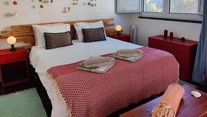 A neatly made bed with a red and white patterned bedspread, two pillows, and a branch with seashells on the wall. There are two bedside tables with lamps, a red nightstand, and a window with a view of palm trees.