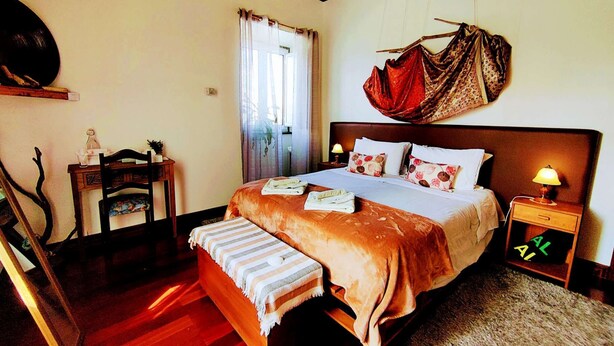 A bedroom with a wooden bed, bedside tables, a chair, a window with curtains, and a wall-mounted artwork. - Two-level marvel in exotic fruit orchard (Ribeira Grande)