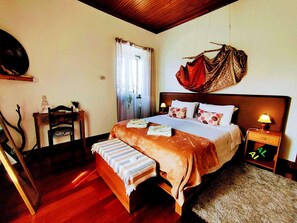 A bedroom with a wooden bed, bedside tables, a chair, a window with curtains, and a wall-mounted artwork. - Two-level marvel in exotic fruit orchard (Ribeira Grande)
