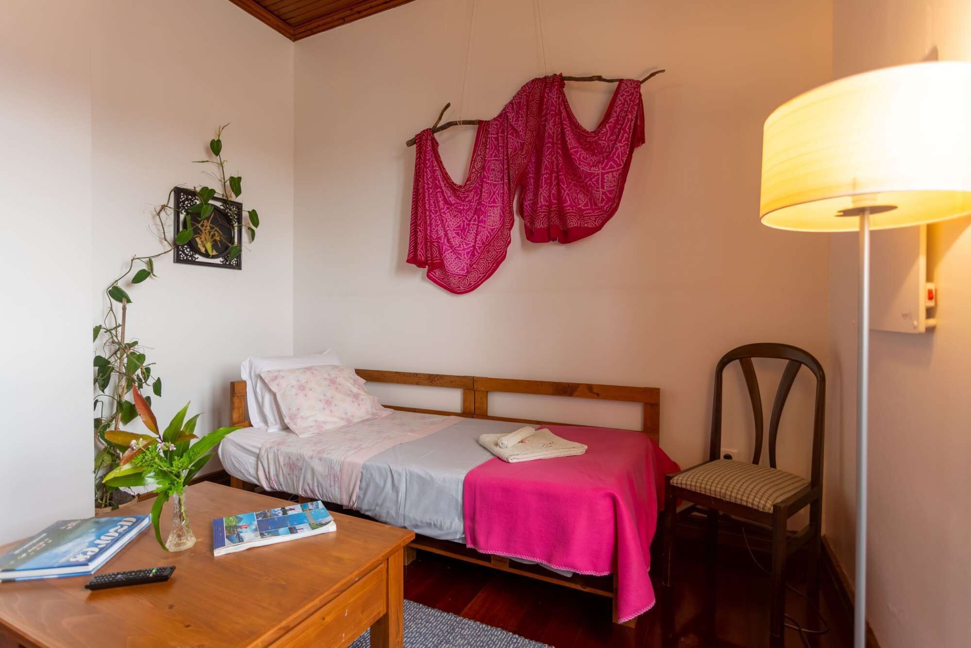 A bedroom with a bed, a desk with a remote control, a lamp, and a chair.