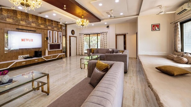 A modern living room with a large TV displaying 'NETFLIX', a sofa, a glass coffee table, and a chandelier. - Geeta Bhawan (Karjat)