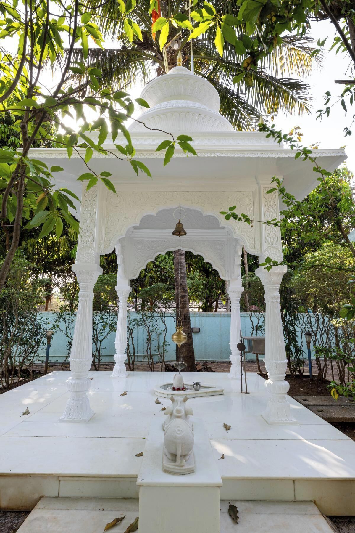 Un belvédère blanc orné de sculptures complexes, entouré d’une végétation luxuriante et adossé à un mur bleu.