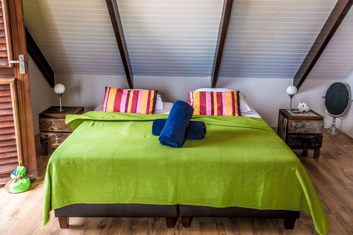 A bedroom with a green bedspread, striped pillows, and a rolled-up blue towel. There are wooden nightstands with lamps and a small round mirror.