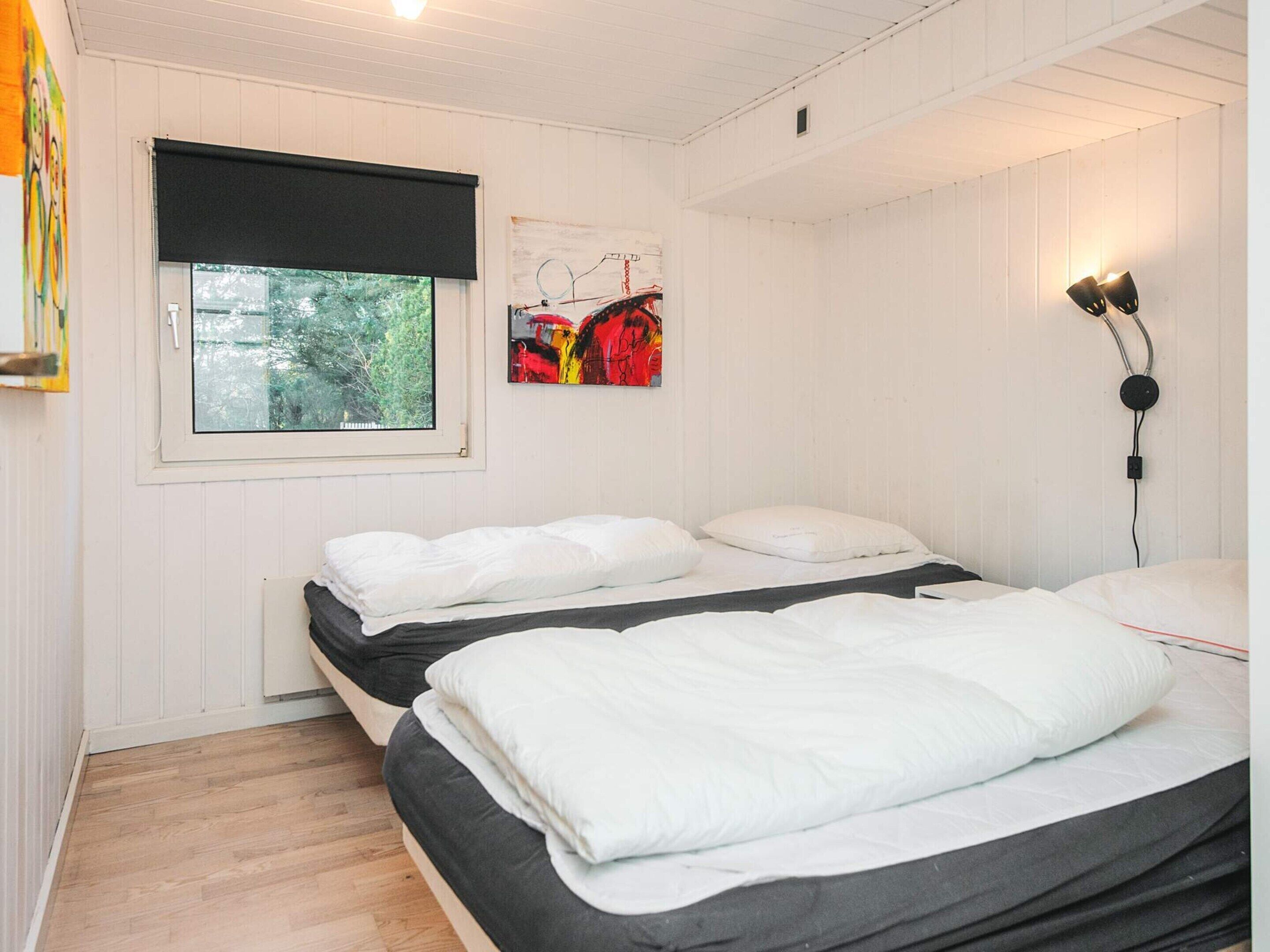 Un dormitorio con dos camas, una ventana con persiana negra, una lámpara de pared y un cuadro.