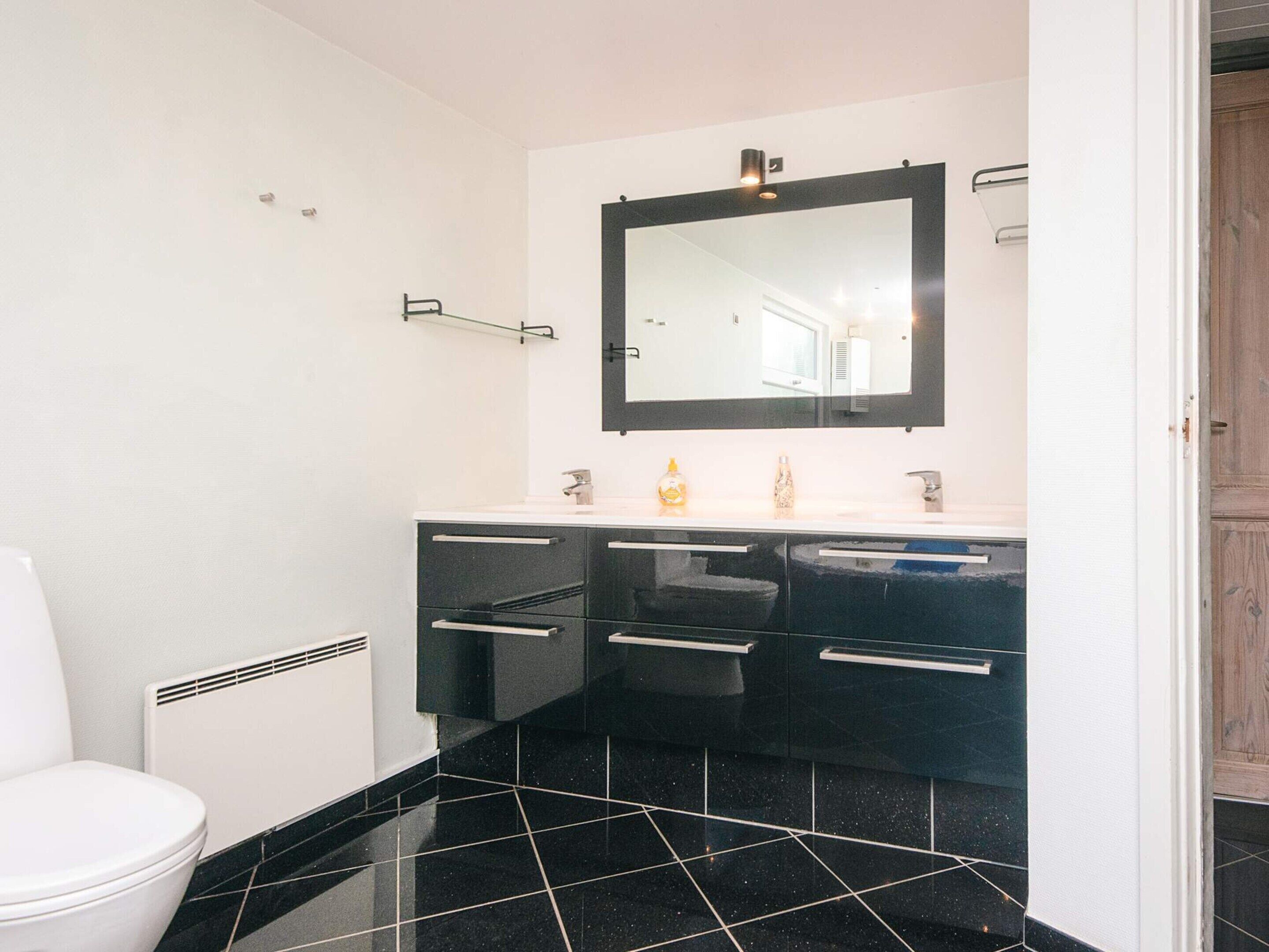 A modern bathroom with a black tiled floor, a white toilet, a black vanity with drawers, a large mirror, and a small shelf with toiletries.