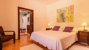 Un dormitorio con una cama grande, dos mesitas de noche con lámparas, una silla, una puerta que da a otra habitación y un cuadro en la pared.