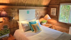 A cozy wooden cabin bedroom with a bed, pillows, bedside lamps, and a window with a view of greenery.