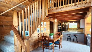 A wooden cabin interior with a dining area, kitchen, and a staircase leading to an upper level.