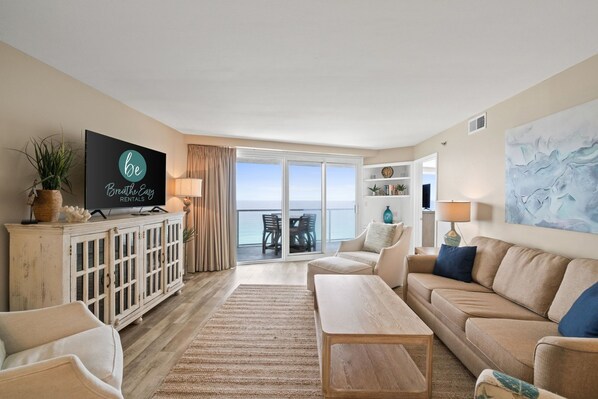 A spacious living room with a large flat-screen TV, a sofa, a coffee table, and a view of the ocean through large sliding doors.