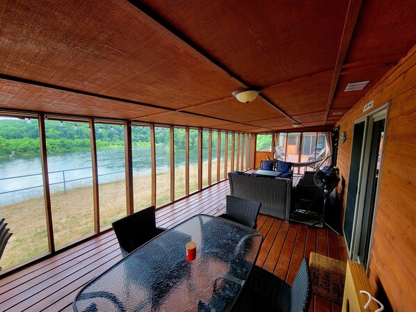 Terraza exterior cubierta con mesa de vidrio y mobiliario de mimbre, con vista al río.