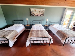 Una habitación con tres camas individuales, cada una con cabecera de madera y colcha blanca. Hay dos mesitas de noche con lámparas, y un cuadro en la pared sobre las camas.