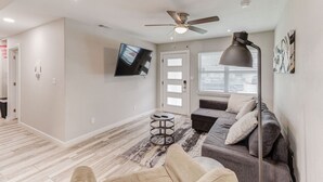 Ein modernes Wohnzimmer mit Deckenventilator, einem flachbildfernseher an der Wand, einem grauen Sofa mit Kissen und einem gemusterten Teppich.