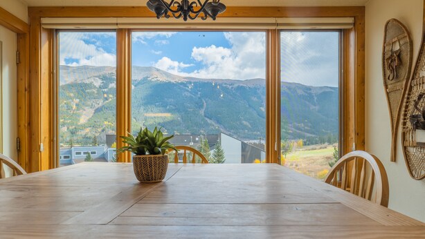 Una mesa de comedor de madera con sillas, una planta en maceta y vistas a montañas a través de amplios ventanales.