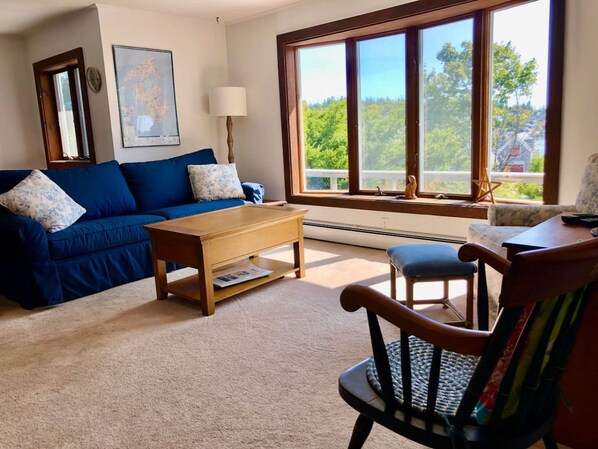 Un salón con un sofá azul, una mesa de centro de madera, una lámpara y vistas a árboles a través de amplios ventanales.