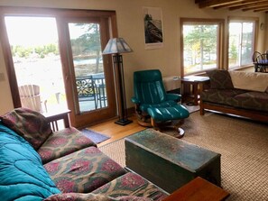 A cozy living room with a sofa, a teal armchair, and a small table. There is a floor lamp and a window with a view of the outdoors. - Dyers Hideout Cottage Beautiful Dyers Island (Vinalhaven)