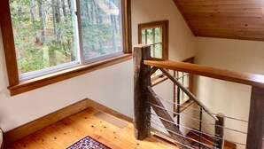 A wooden staircase with a metal railing leading to a second floor, large windows offering a view of a forest, and a wooden floor. - Dyers Hideout Cottage Beautiful Dyers Island (Vinalhaven)