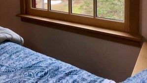 A bedroom with a large window, a bed with a blue patterned bedspread, and a view of trees and a building outside. - Dyers Hideout Cottage Beautiful Dyers Island (Vinalhaven)