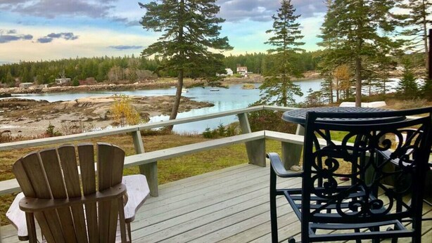 A wooden deck with two Adirondack chairs and a table, overlooking a river and forested area. - Dyers Hideout Cottage Beautiful Dyers Island (Vinalhaven)