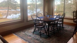 A dining area with a table and chairs, large windows with a view of a lake and trees, and a patterned rug on the floor. - Dyers Hideout Cottage Beautiful Dyers Island (Vinalhaven)