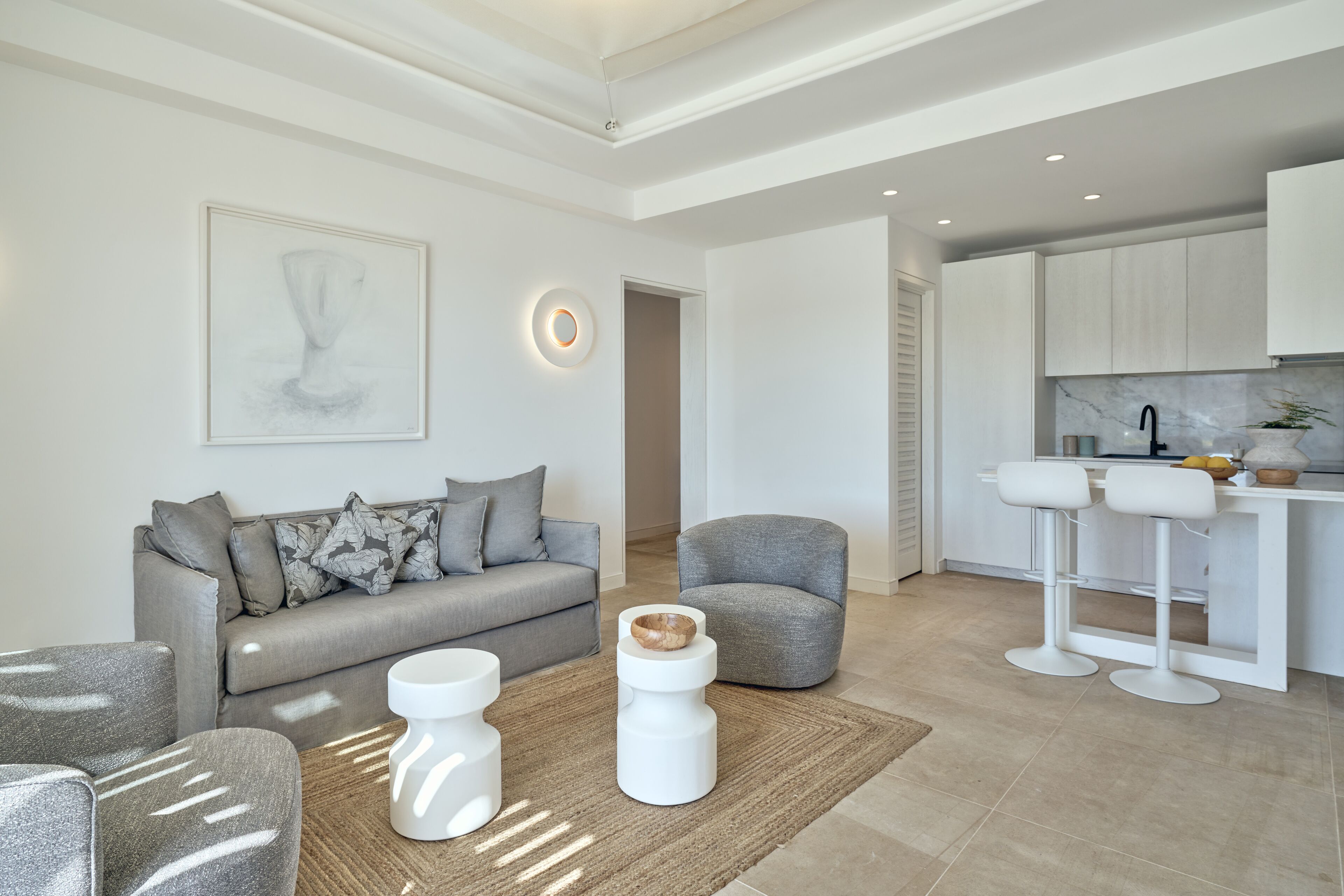 A modern living room with a grey sofa, a white coffee table, and a grey armchair. There is a kitchen area with white cabinets and a marble backsplash.