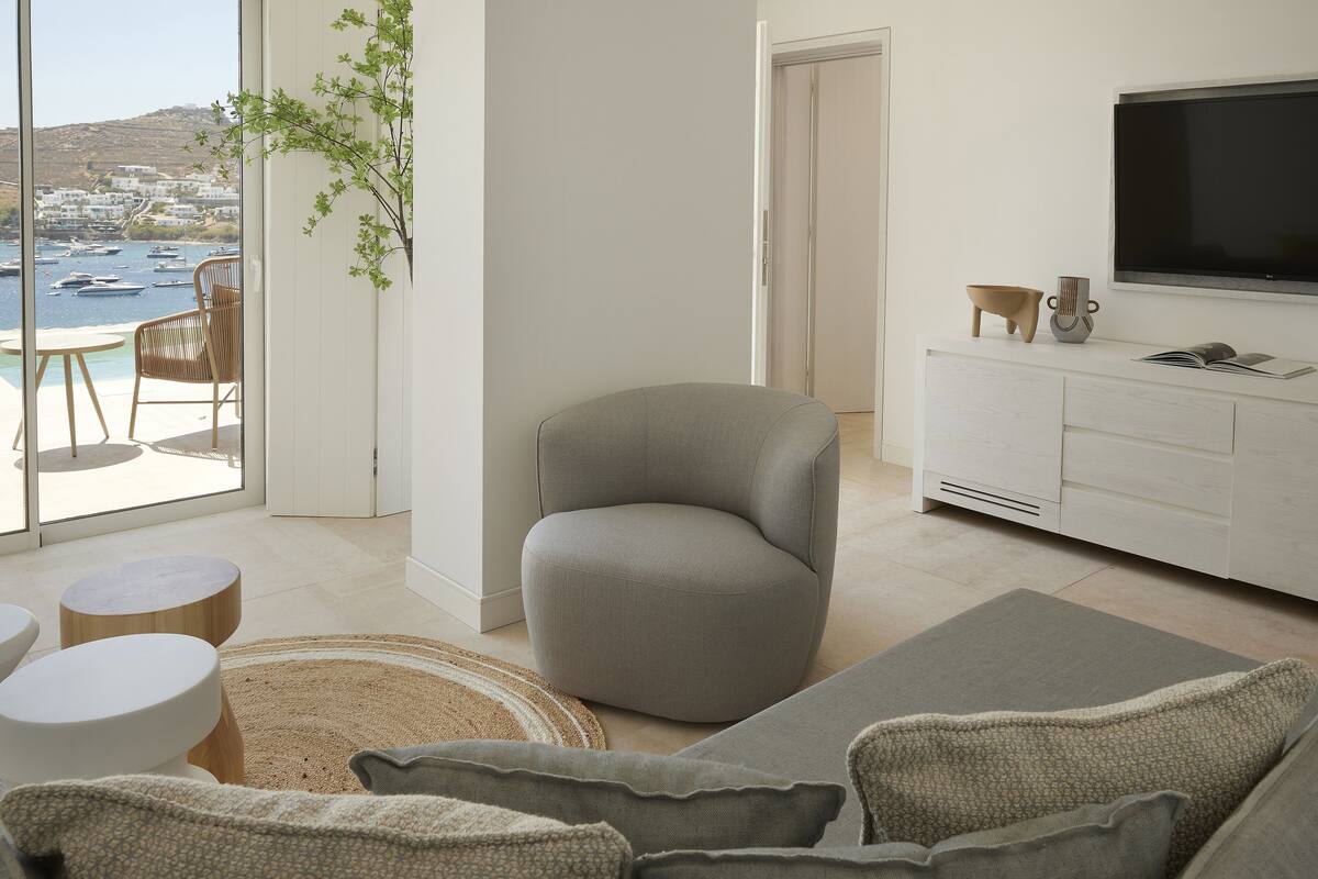Un salon moderne avec un canapé gris, un meuble TV blanc et une vue sur les bateaux qui naviguent, visible à travers de grandes fenêtres.