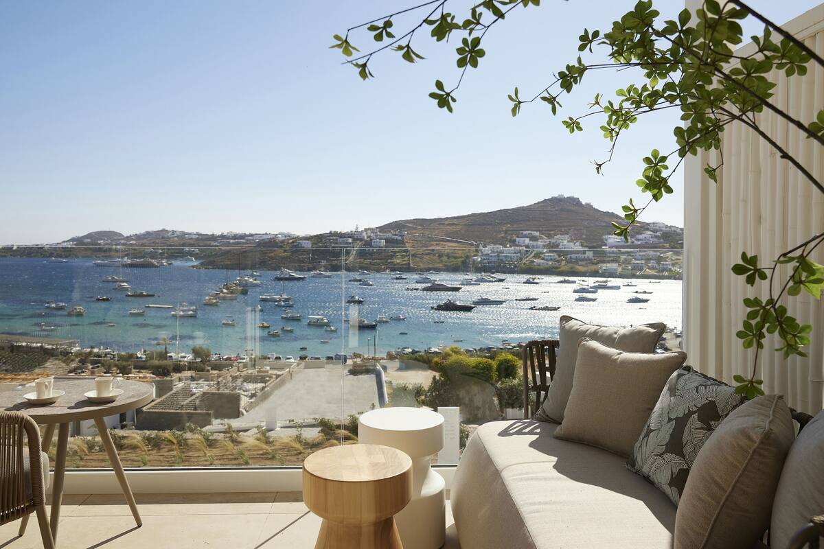 Un balcon donnant sur la mer, avec vue sur les bateaux, une table avec des tasses et un canapé avec des coussins.
