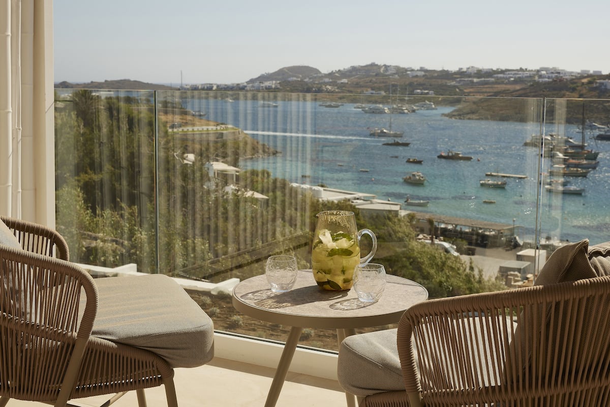 Un balcon avec des fauteuils en osier et une table, donnant sur une zone côtière où l’on peut apercevoir des bateaux et des bâtiments.