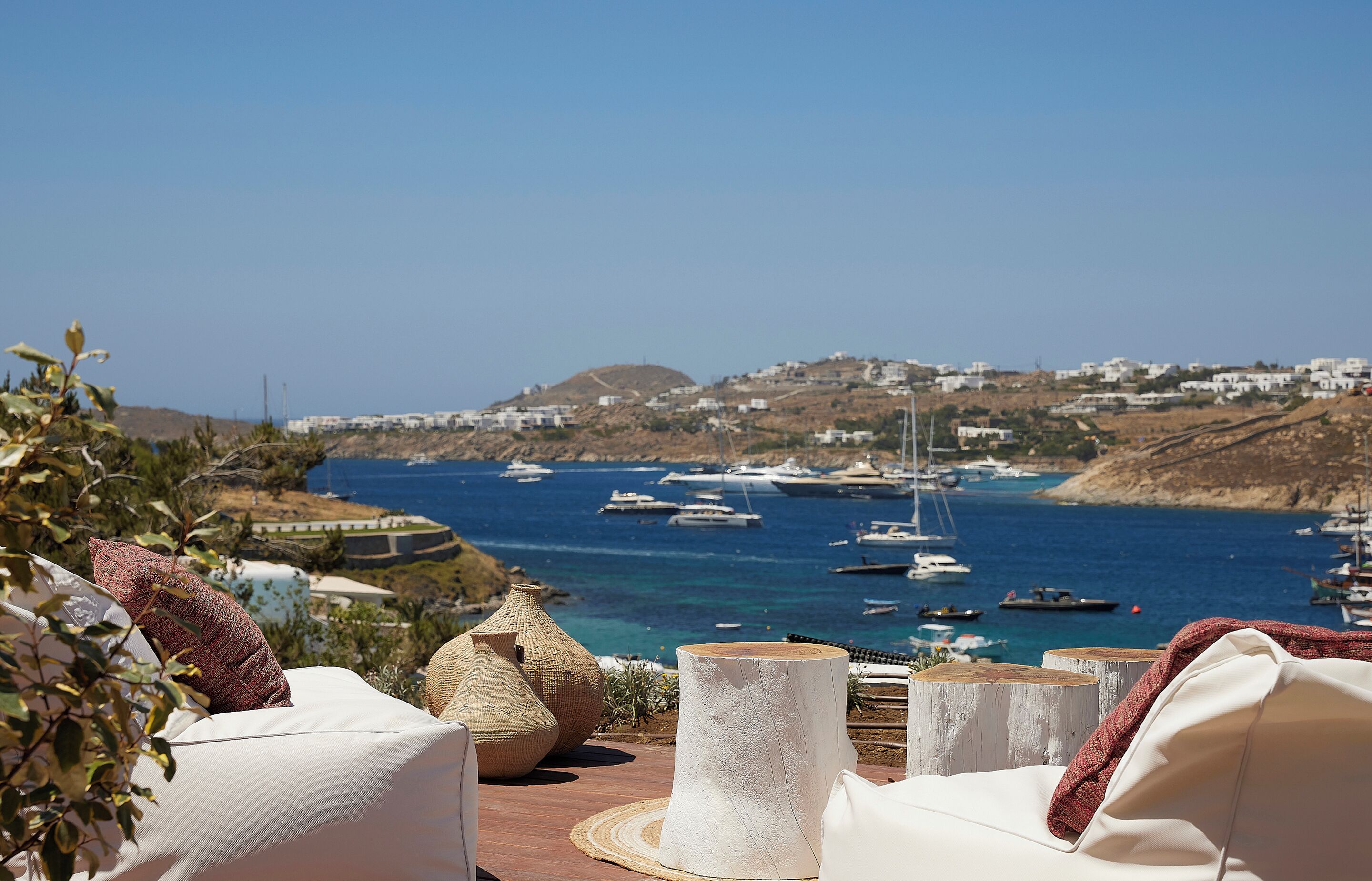 A terrace with white lounge chairs, a woven basket, and a rustic wooden table. In the background, there is a view of a coastal town with boats and white buildings.