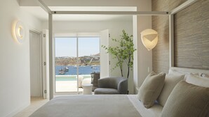 A modern bedroom with a large bed, a view of boats and a coastal landscape through a sliding door, and a small table with a chair.