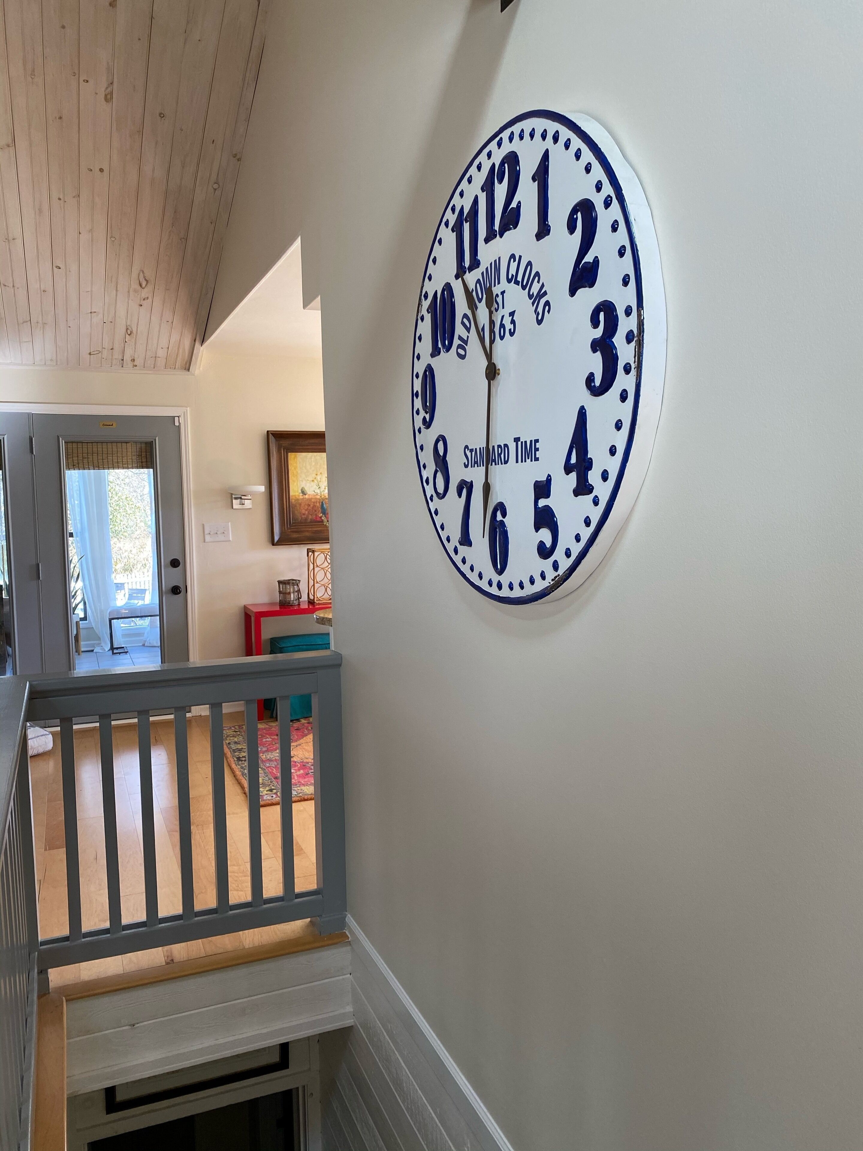 A wall-mounted clock with a classic design, a staircase with a wooden railing, and a glimpse of a room with a window and a door.