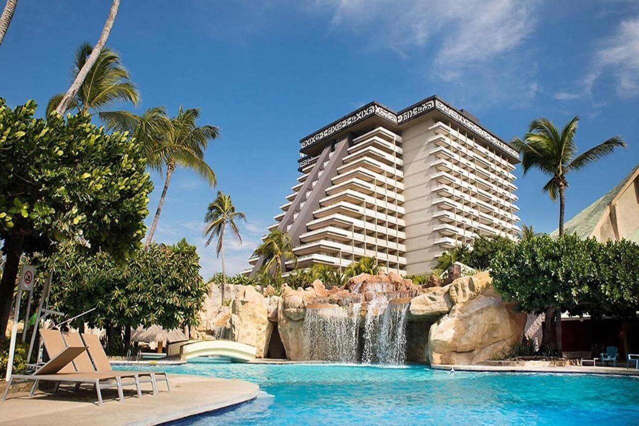 A hotel with a pool, palm trees, and a waterfall.
