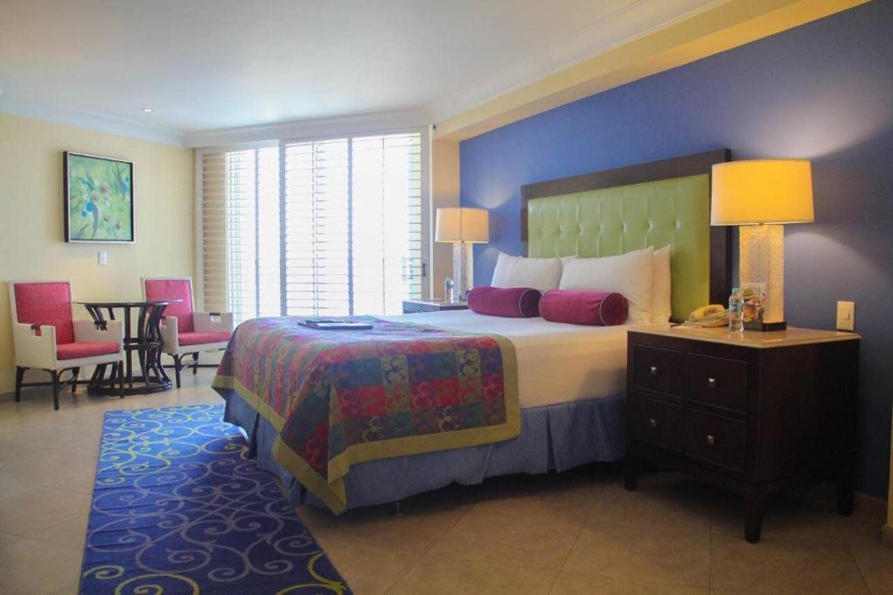 A hotel room with a large bed, two bedside lamps, a small desk with a chair, and a window with blinds.