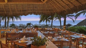 An outdoor dining area with tables and chairs, a thatched roof, and a view of the ocean. - Princess Mundo Imperial (Acapulco)