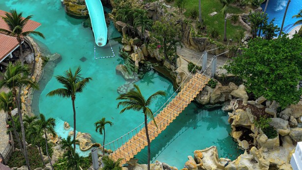 A rope bridge over a turquoise pool with palm trees and rocky formations. - Princess Mundo Imperial (Acapulco)