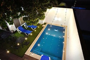 Ein rechteckiger Swimmingpool mit blauer Innenseite, umgeben von einer kleinen Rasenfläche und einigen Liegestühlen.